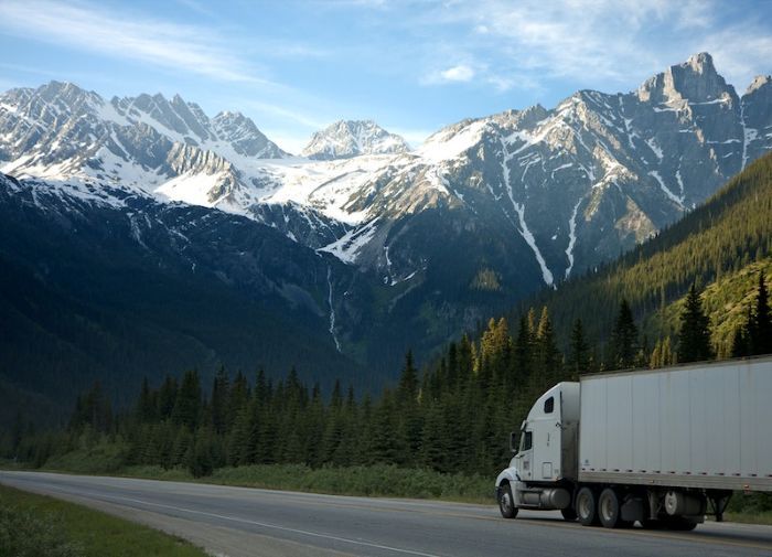White long distance moving truck driving past snow-capped mountains on a cross country highway.